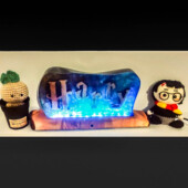 Quand la lumière réveille la magie. Lampe artisanale “Harry” en bois et résine époxy bleue, éclairée par LED, posée entre deux figurines décoratives.